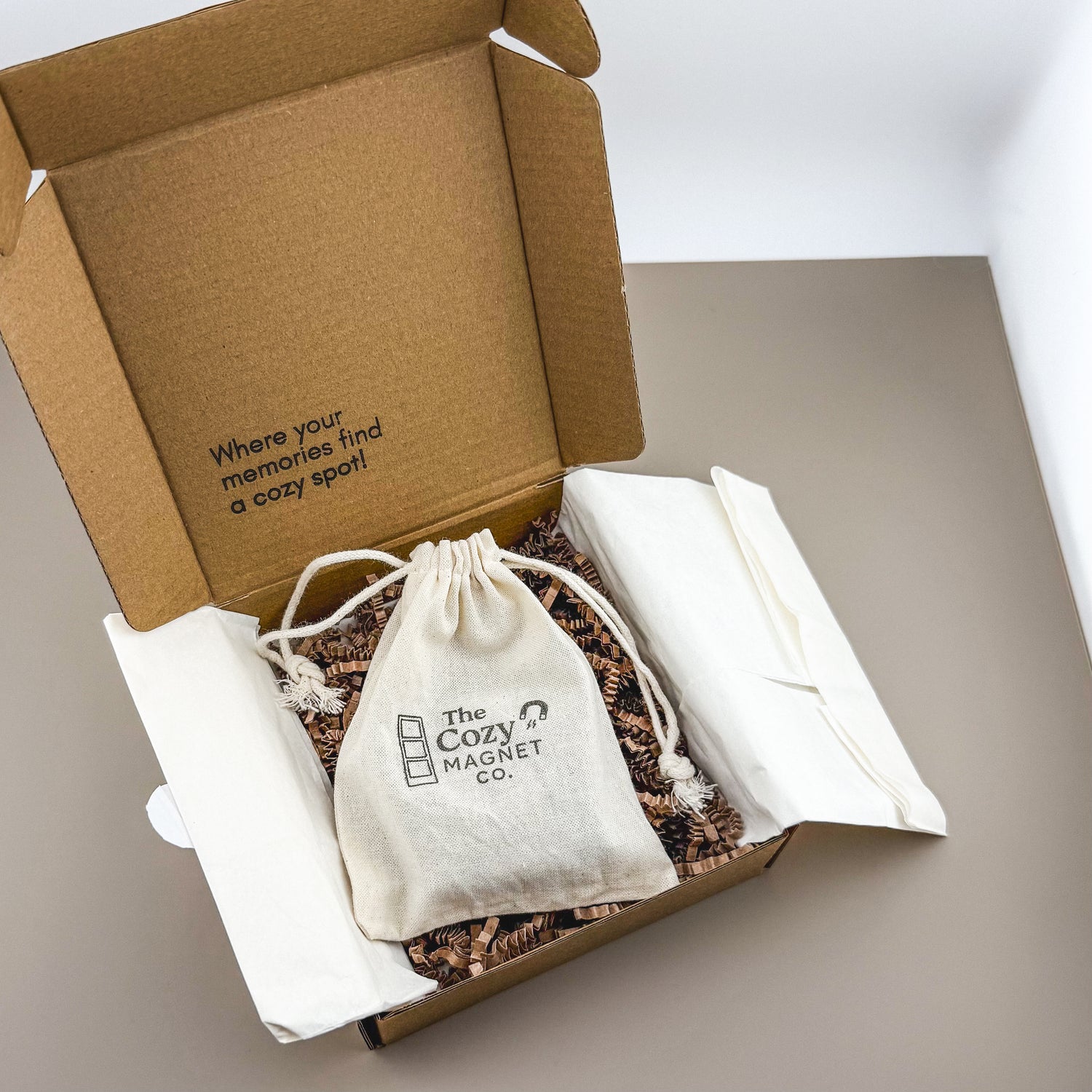 Open cardboard box with 'The Cozy Magnet Co.' product inside on a neutral background