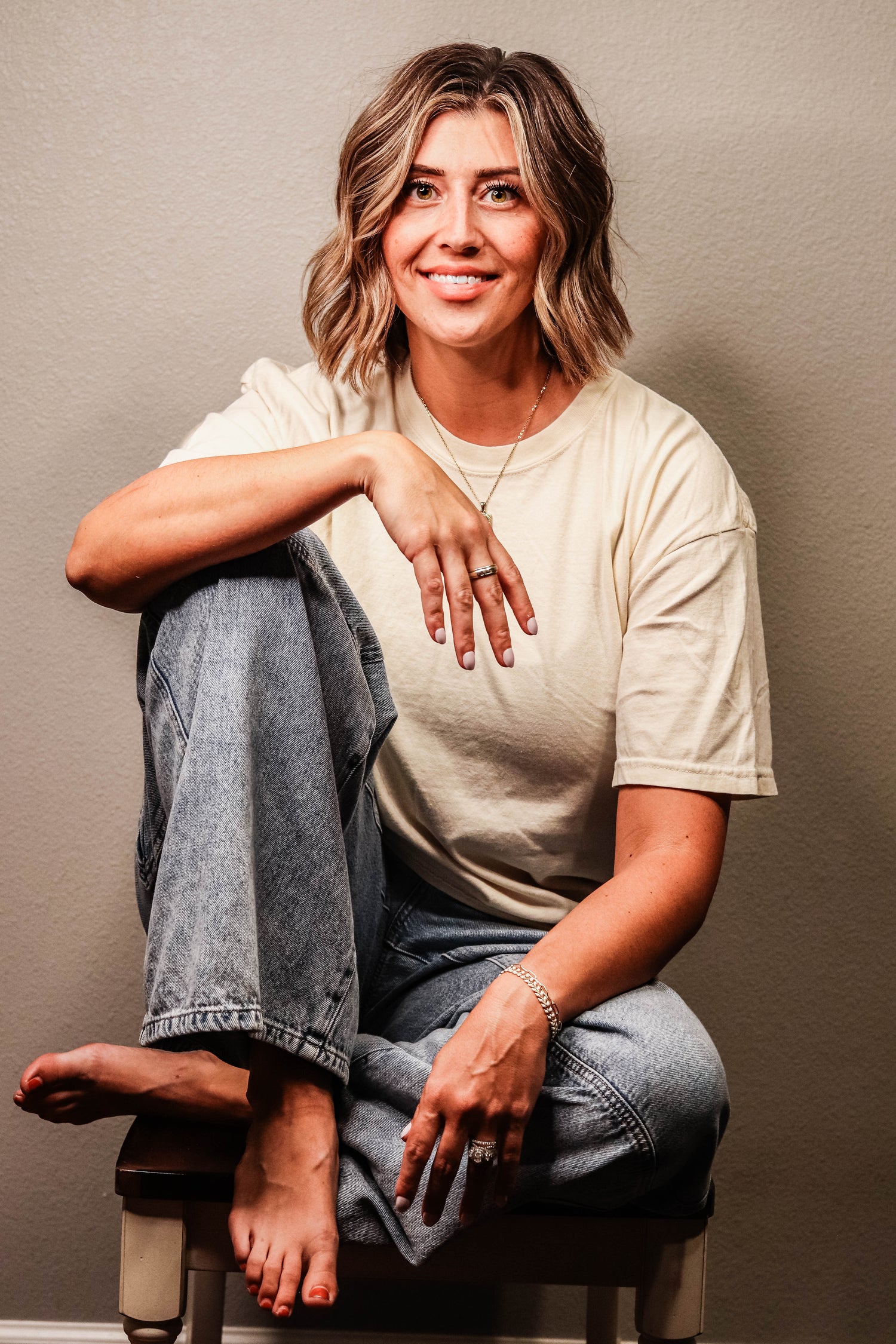 A woman with shoulder-length blonde-highlighted hair sits barefoot on a stool against a plain beige wall. She is wearing a cream-colored t-shirt and light-wash blue jeans, with her right arm resting on her knee.